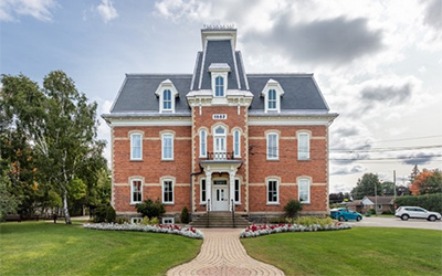 Une maison en briques de deux étages dotée d’un toit gris ardoise, d’un balcon central au deuxième étage et de nombreuses fenêtres symétriques bordées de blanc se trouve au centre de la photo, entourée d’un ciel bleu et d’une pelouse verte