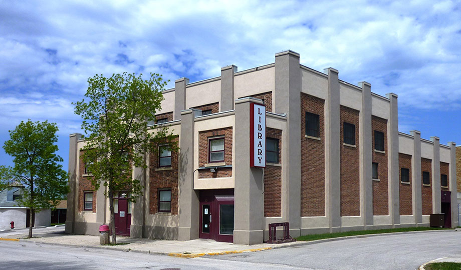 Vue de face de la bibliothèque régionale de The Pas, un bâtiment en briques rouges et beiges bordé de deux arbres à l’entrée.