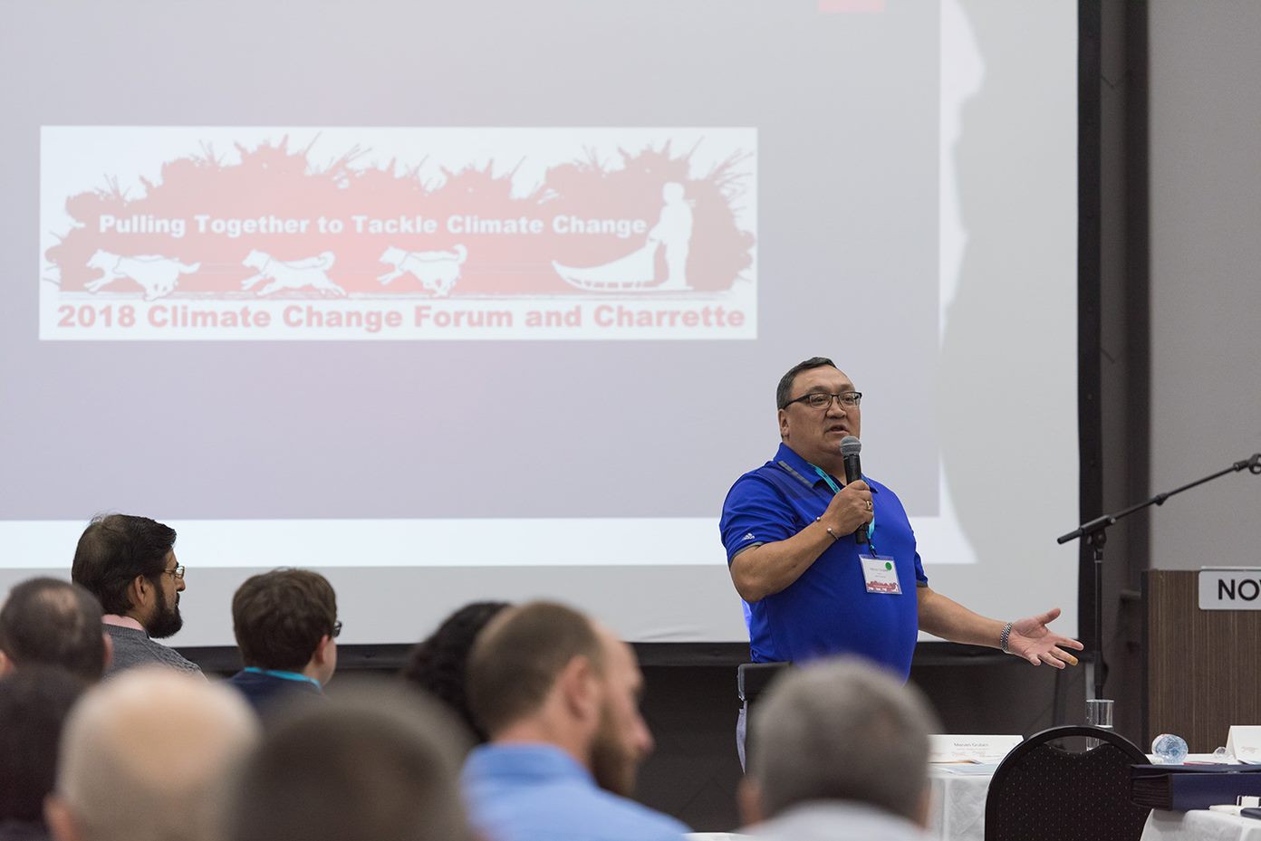 Un homme debout en chemise bleue tient un microphone pendant qu'il présente aux participants assis au forum, projeté derrière lui un écran disant «Forum et Charrette sur le changement climatique 2018»