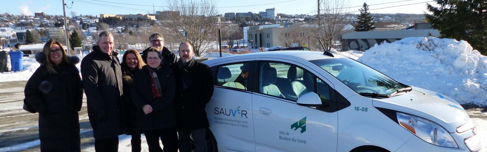 Des représentants des municipalités sourient à la caméra devant l’un des véhicules électriques, par une journée d’hiver ensoleillée.