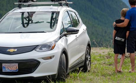 An electric vehicle, bearing two mountain bikes on the roof rack, parked atop a lookout beside a young couple embracing and taking in a stunning landscape of mountains of trees