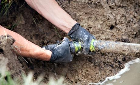 Two gloved hands grip an exposed underground pipe covered in mud.